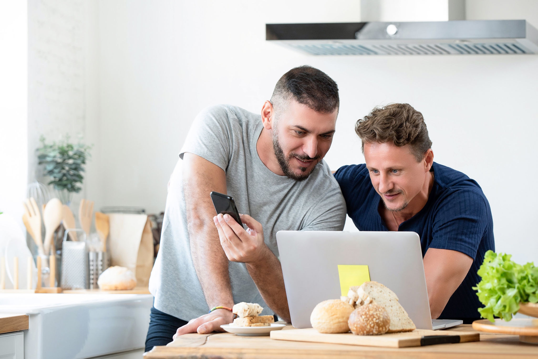 2 people looking at a laptop in the Kitchen one holds a smartphone