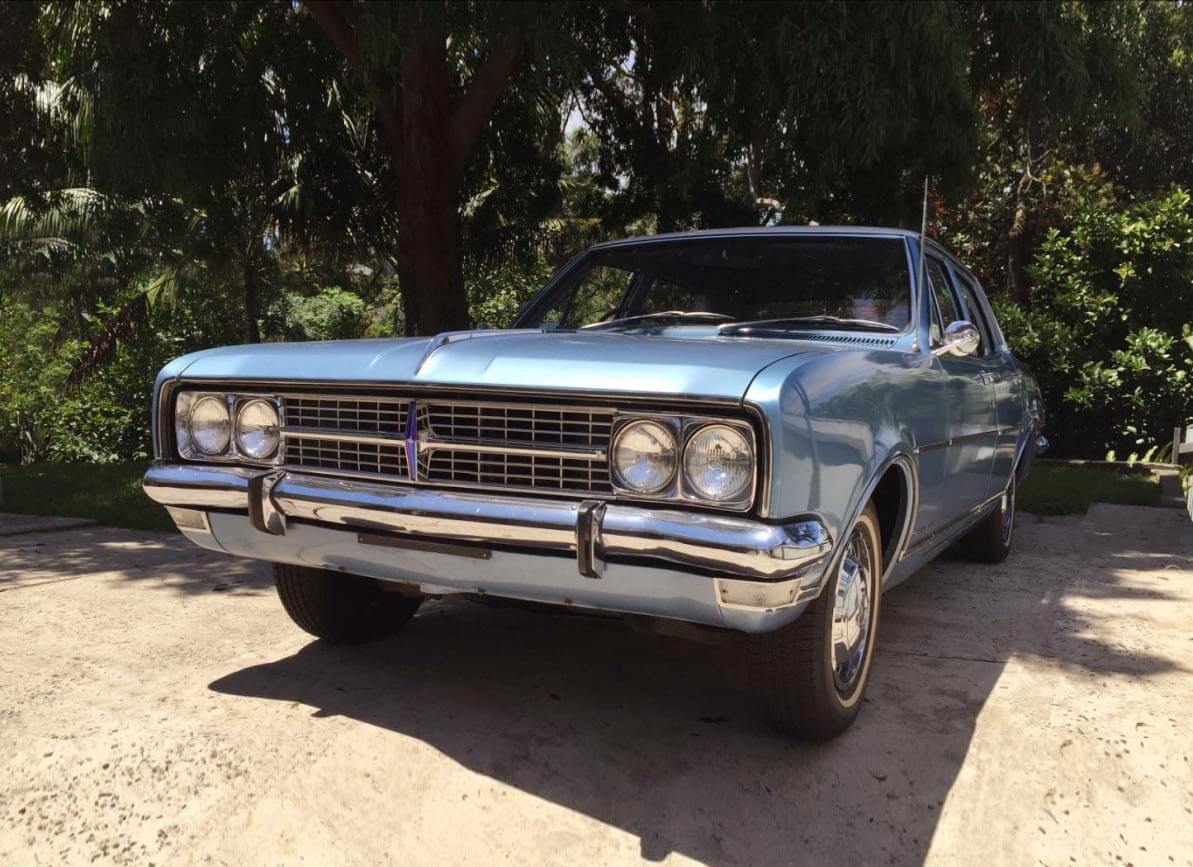 A silvery blue classic car parked in front of trees