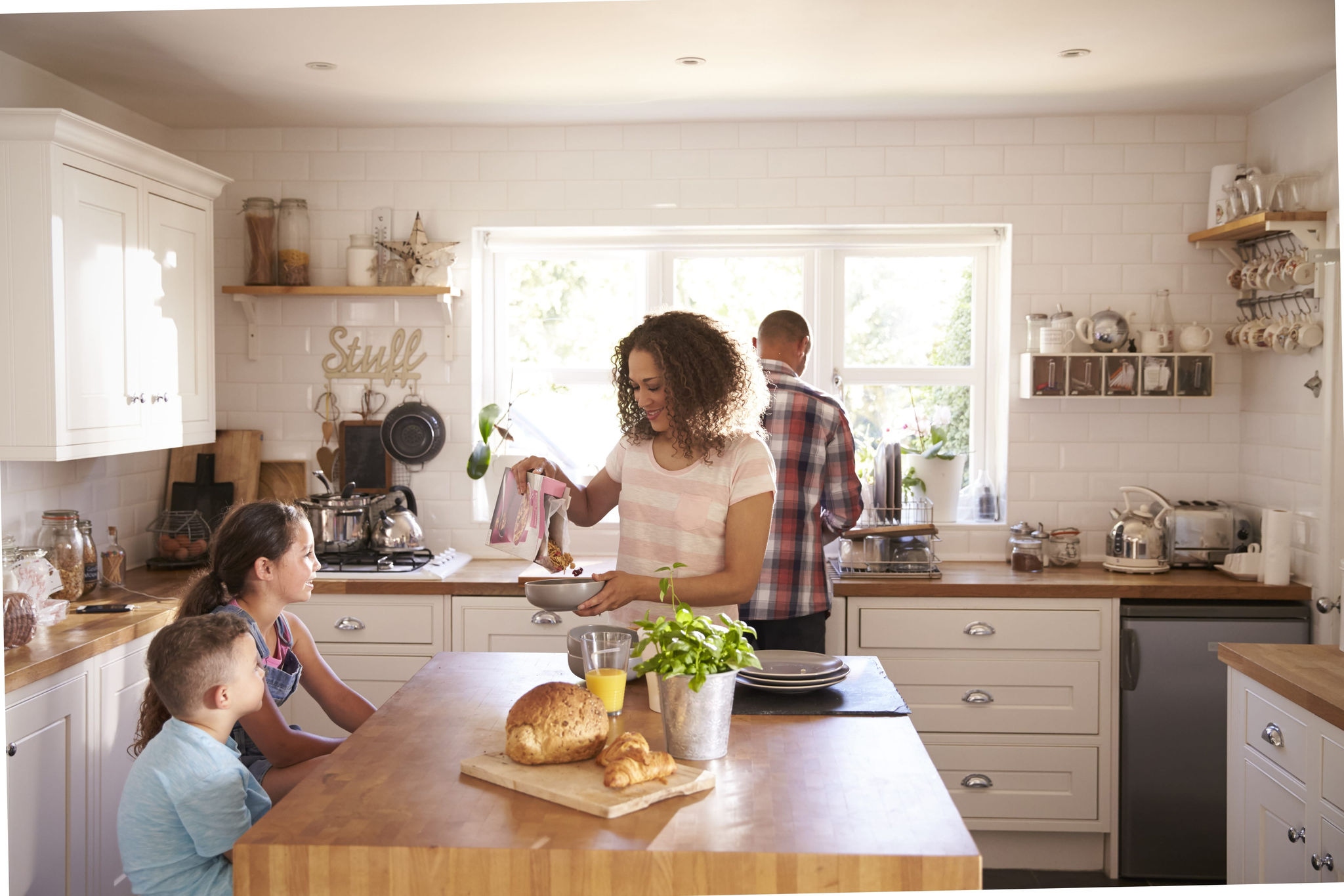 Parent pouring cereal, other parent at sink. children at table