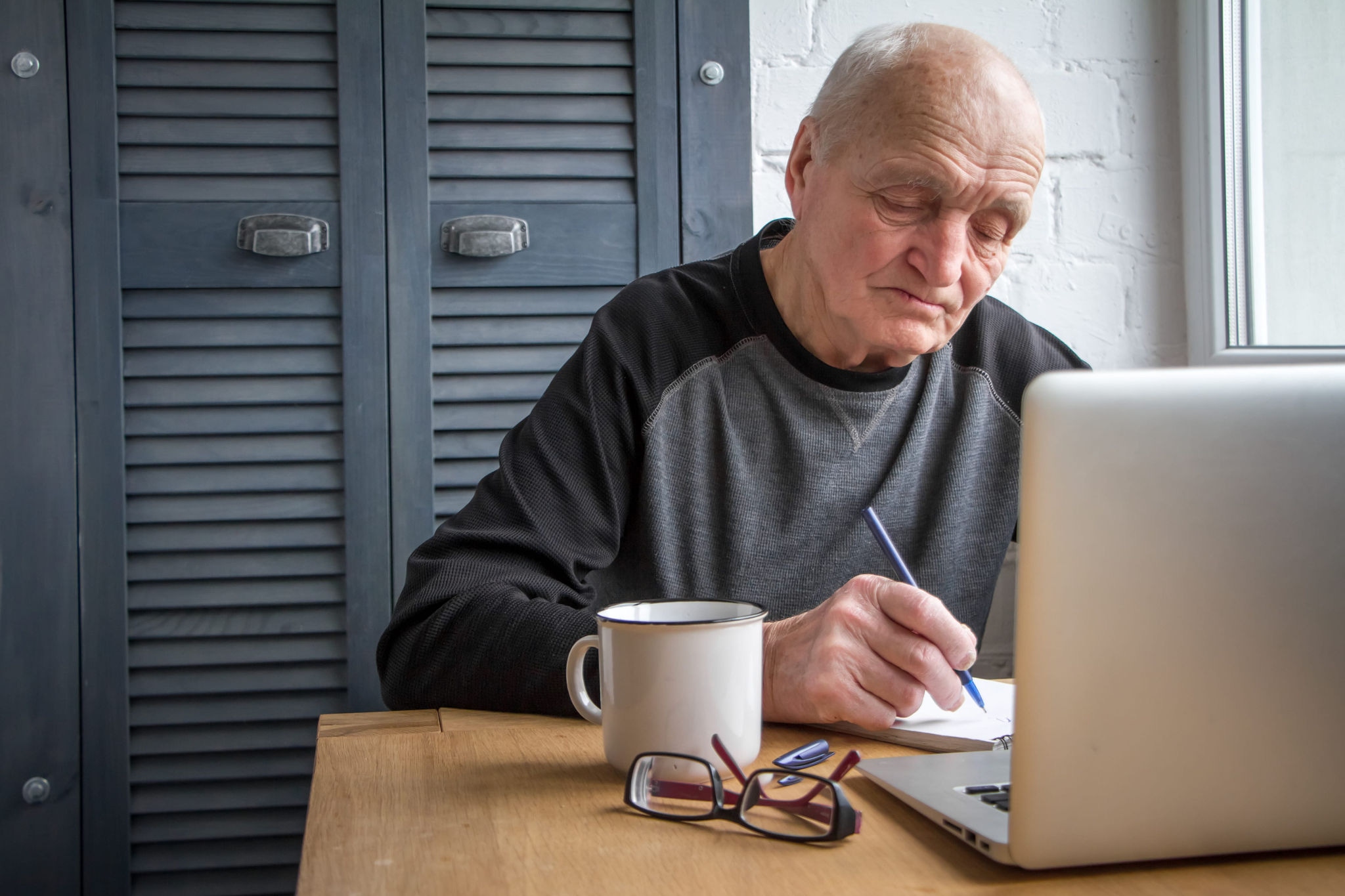 Person sitting in front of laptop, writing on a notepad with glasses and a coffee cup on the table.